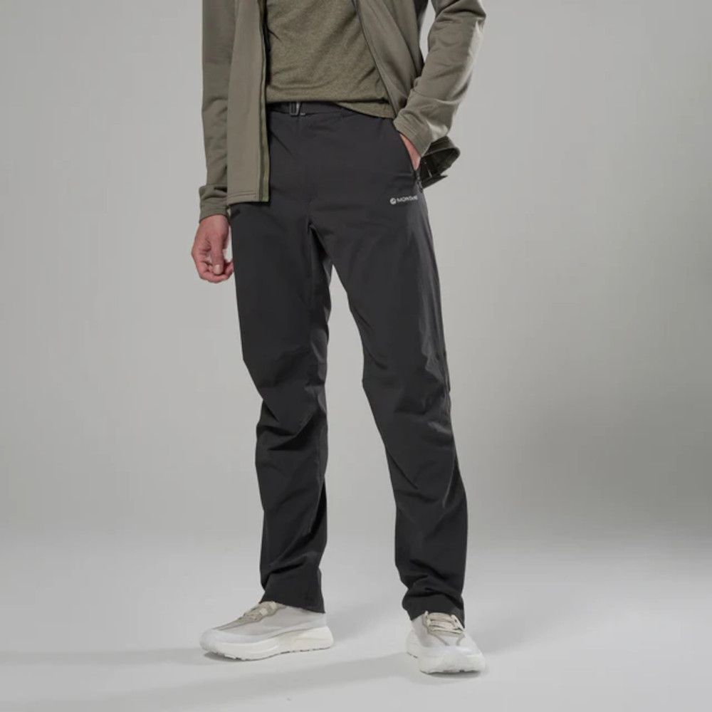 Person standing in dark gray outdoor hiking pants with one hand in a pocket, wearing an olive jacket and white sneakers.