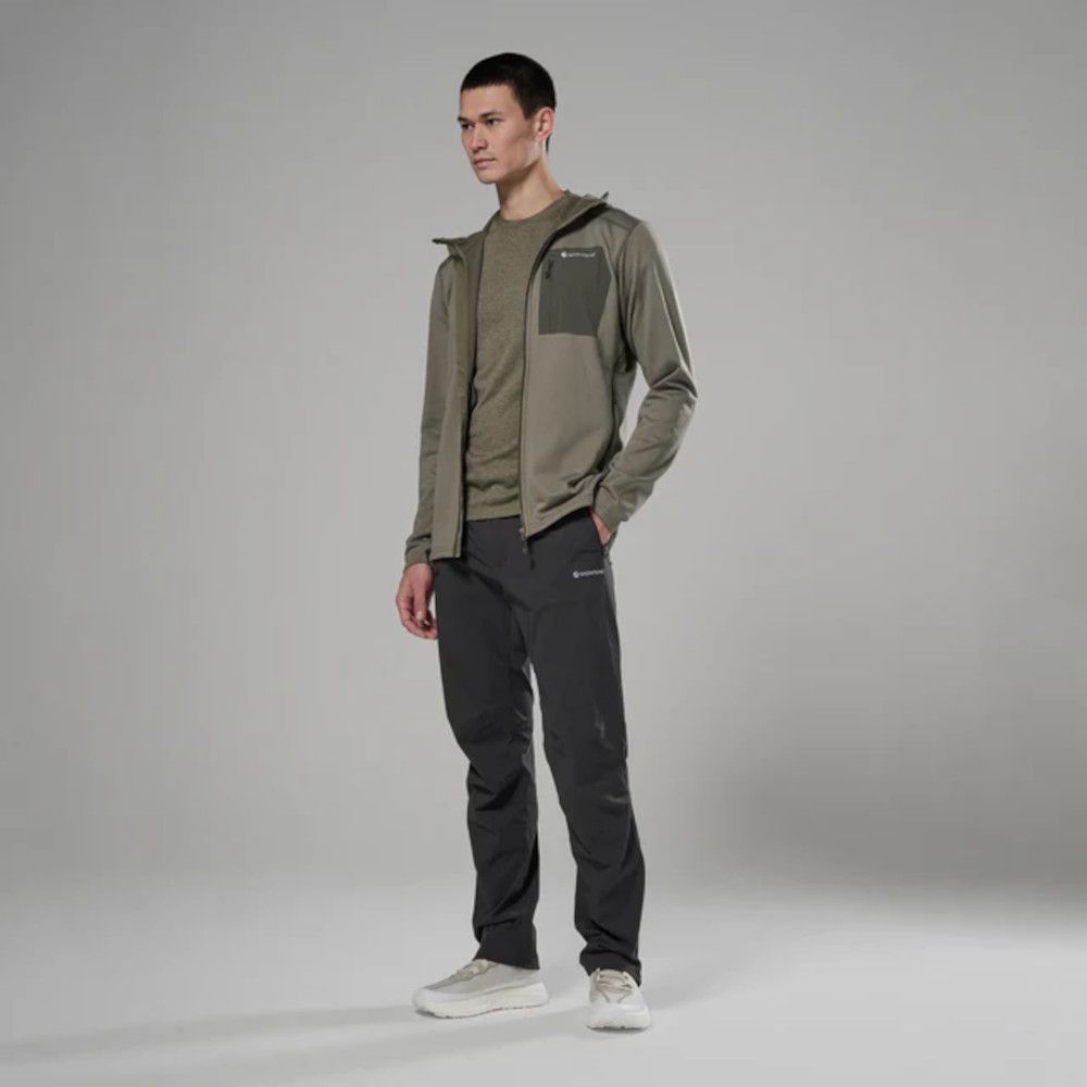 Man modeling an olive windbreaker over a greenish shirt with dark gray outdoor pants and white sneakers in a studio setting.
