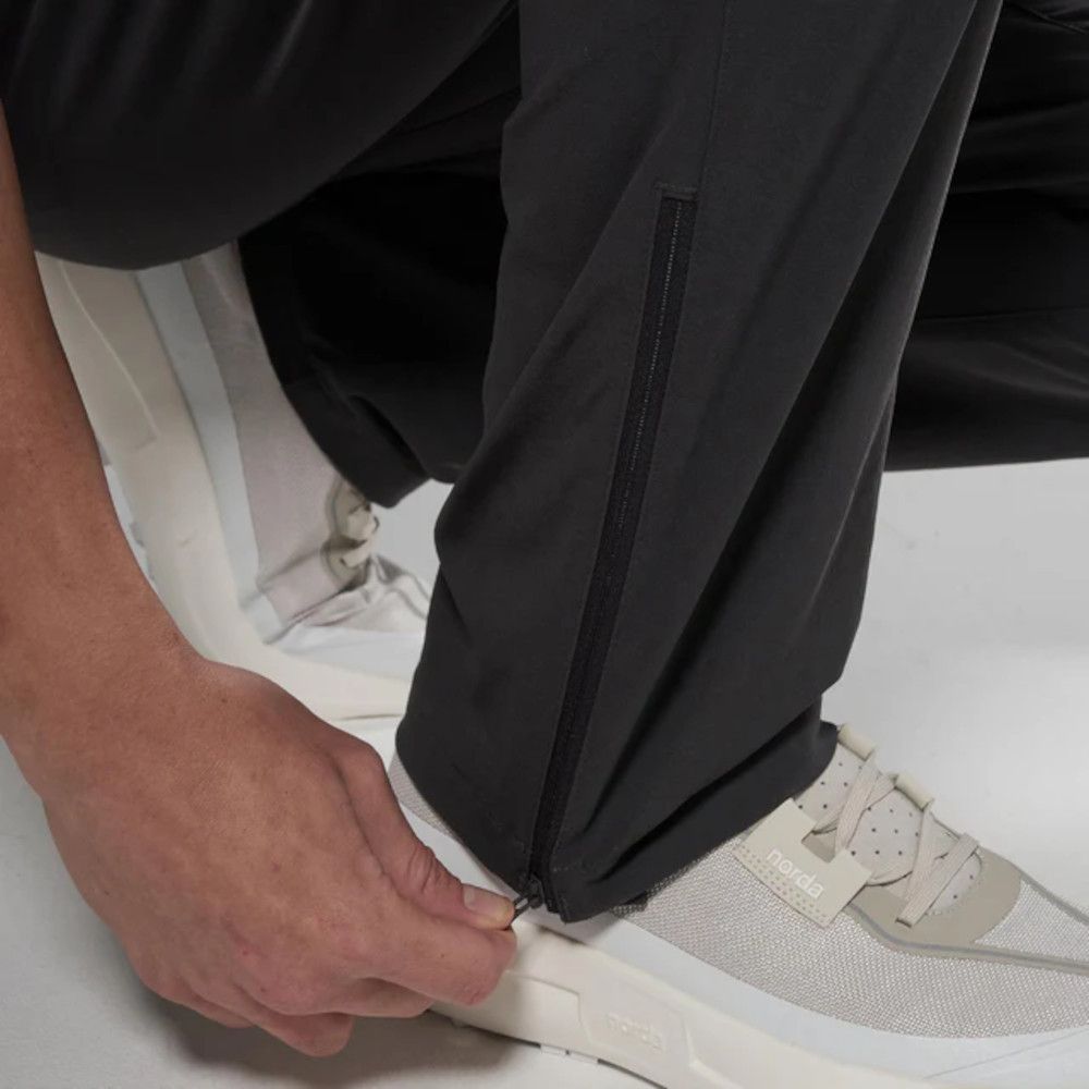 Close-up of a hand zipping the cuff of black pants over a light gray sneaker with a beige sole side view.