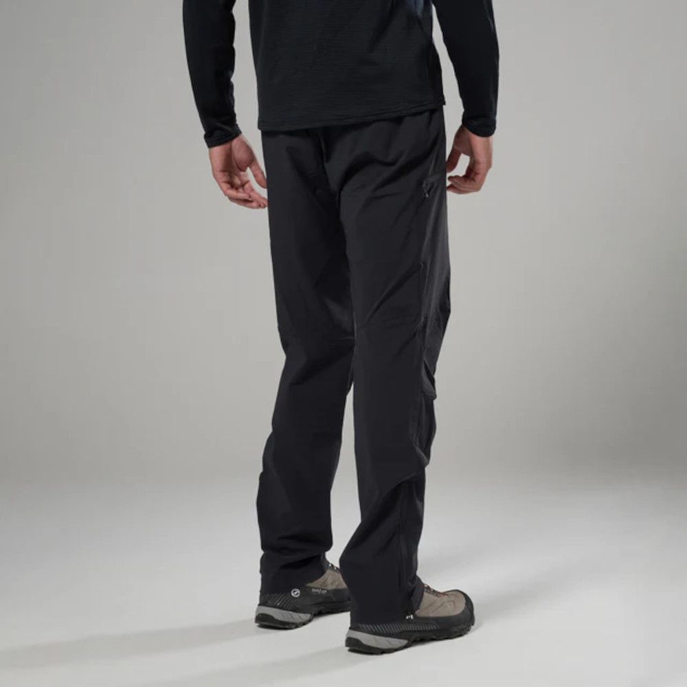 Back view of a person wearing black hiking pants and gray-brown hiking shoes against a light gray background.