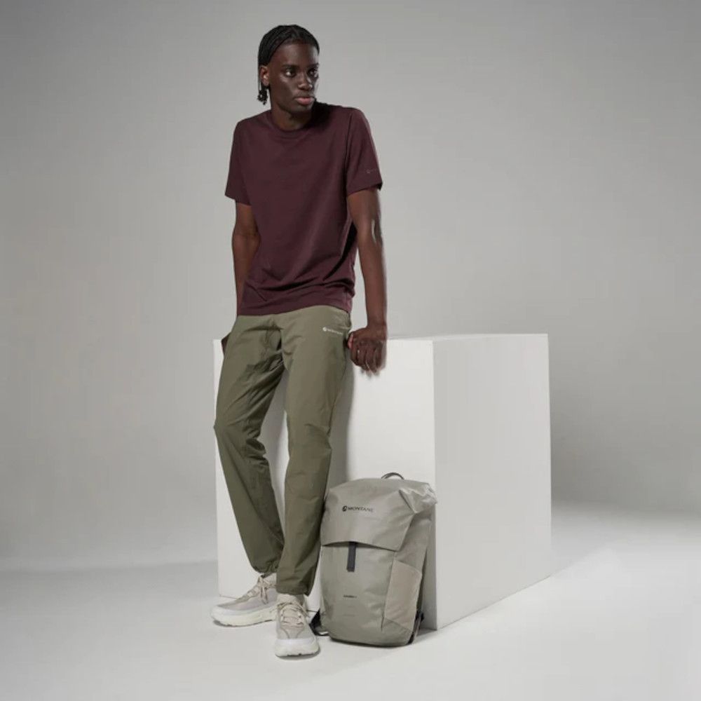 Young man leaning against a white block, wearing a burgundy T-shirt, olive pants, beige sneakers, with a gray backpack beside him on the floor.