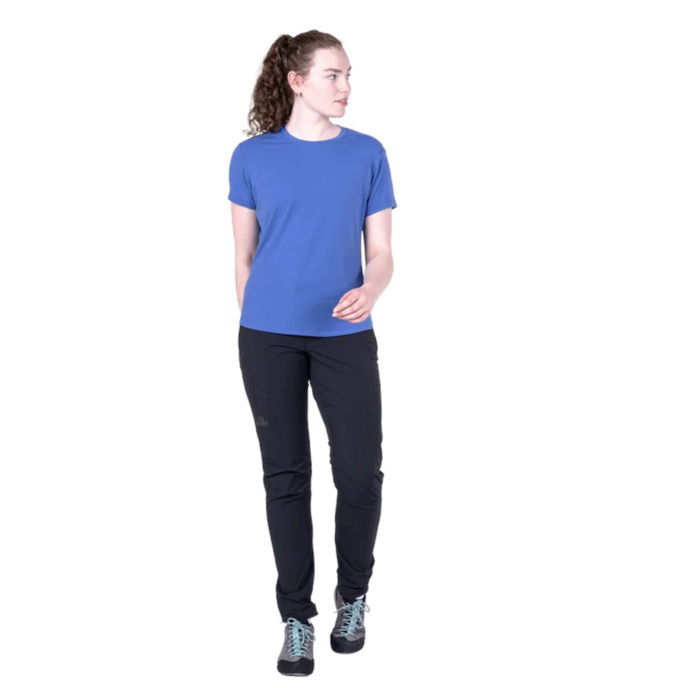 1_43c614ff-305c-487c-b528-744a22aa94cf Woman standing in a blue short-sleeve t-shirt and black hiking pants with teal-laced boots, looking to her left.