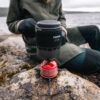 1_43c614ff-305c-487c-b528-744a22aa94cf Person on rocky shore using a Primus Lite XL camping stove with a red fuel canister beside it, preparing to boil water outdoors.