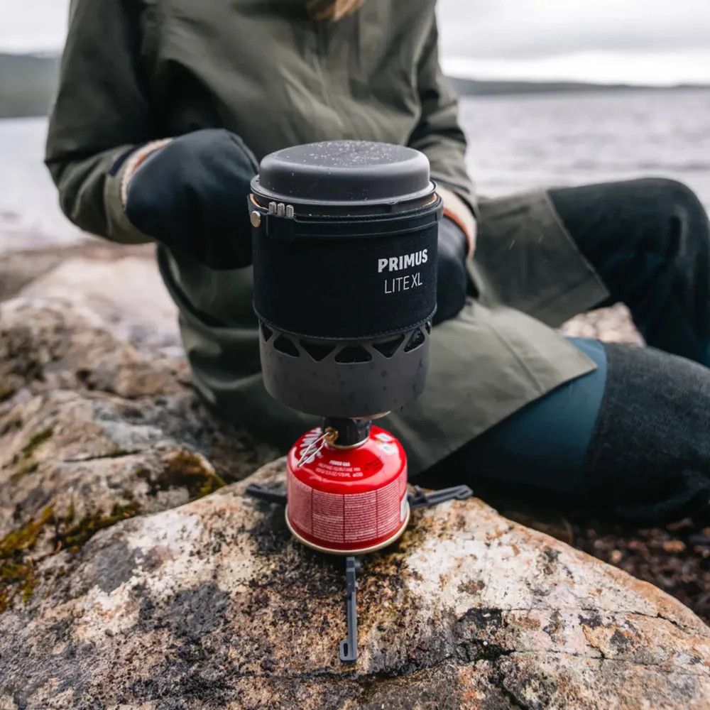 1_43c614ff-305c-487c-b528-744a22aa94cf Person on rocky shore using a Primus Lite XL camping stove with a red fuel canister beside it, preparing to boil water outdoors.