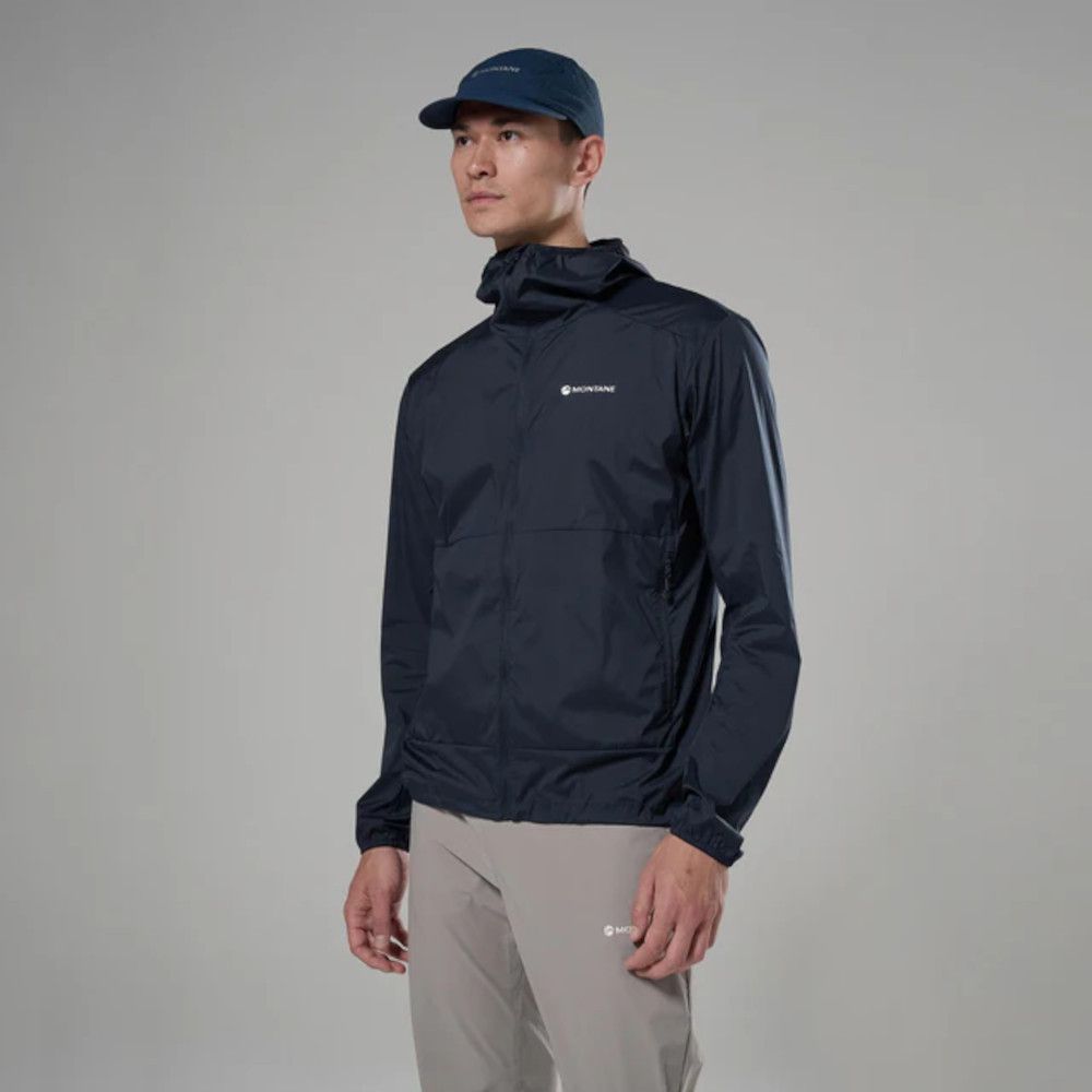 1_43c614ff-305c-487c-b528-744a22aa94cf Man modeling a navy Montane windbreaker with a small logo, wearing a blue cap and light gray pants in a studio setting.