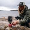 1_43c614ff-305c-487c-b528-744a22aa94cf Woman outdoors by a lake setting up a small camping stove on a rock, using a red gas canister labeled Primus Lite+ with a gray pot above.