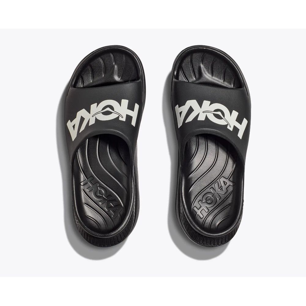 1_43c614ff-305c-487c-b528-744a22aa94cf Pair of black Hoka slide sandals photographed from above, showing a large white logo on each strap on a light background.