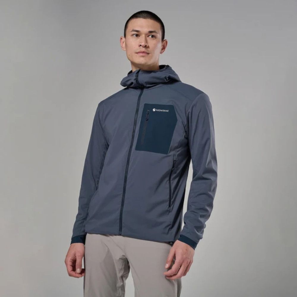Man modeling a gray-blue windbreaker with a dark chest pocket and hood, standing against a neutral backdrop in a studio.