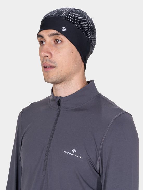 RH-009061_Reflect_Beanie_R954_Black_Reflect-1078 Man wearing a dark knit beanie with a small logo and a gray Ronhill quarter-zip athletic top.